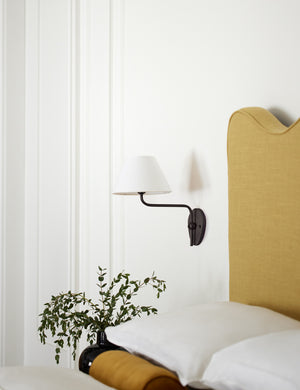 The Magdalene black single sconce is mounted to the right of a golden linen bed on a white wall