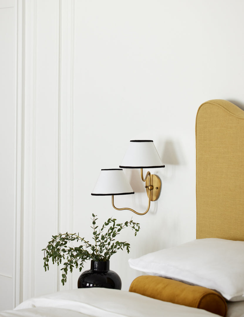 #color::black-trim #finish::brass | The Magdalene brass double sconce is mounted to the right of a golden linen bed on a white wall