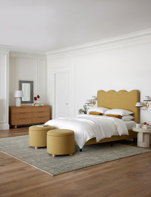 Two kamila golden linen 24 inch ottomans sit at the end of a golden linen framed bed with a scalloped headboard atop a rug