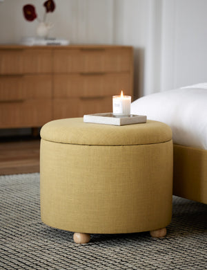 The kamila golden linen 24 inch ottoman sits at the edge of a bed atop a black and white grid patterned rug with a candle atop it
