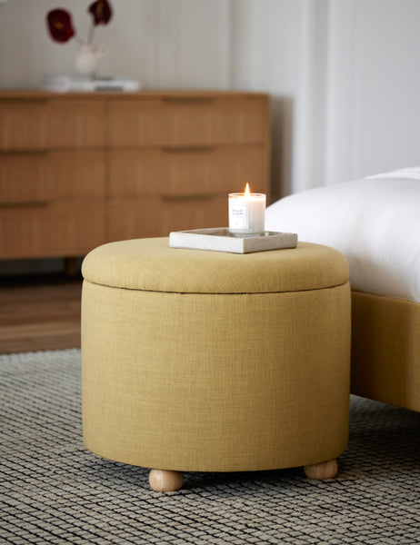 #color::golden-linen #size::24-Dia | The kamila golden linen 24 inch ottoman sits at the edge of a bed atop a black and white grid patterned rug with a candle atop it