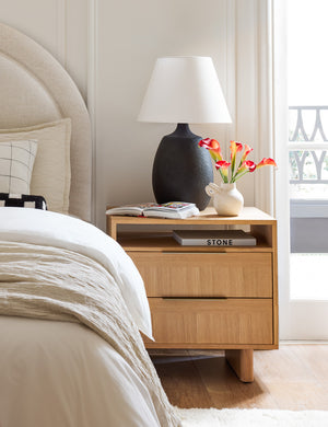 The Pratt black table lamp sits on a wooden nightstand with a book and a white vase next to a linen framed bed