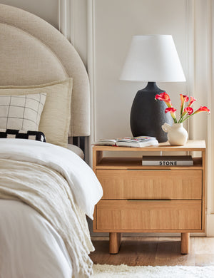 The Hillard white oak veneer nightstand sits next to a bed with an arched linen frame with a black lamp and a white vase