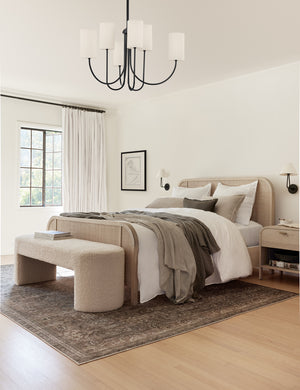 The Mikhail cream boucle foam-padded bench sits in a bedroom at the end of a bed with a natural woven bed frame and underneath a black chandelier pendant.
