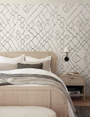 The Moroccan black and ivory Wallpaper Mural by Sarah Sherman Samuel is in a bedroom with a woven cane bed, a light wood nightstand, and a black double armed sconce.