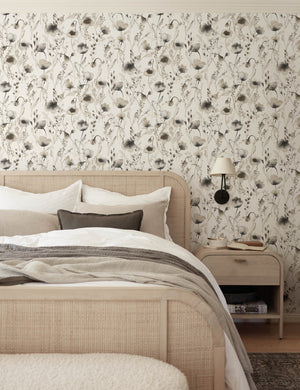 The Scalamandre Lo soft and pastoral brown toned floral wallpaper is in a bedroom with a cane and woven framed bed, gray toned bedding, and a whitewashed wooden nightstand
