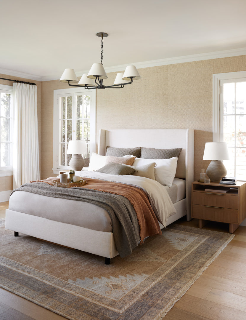 #color::talc-linen #size::cal-king #size::full #size::king #size::queen #size::twin | The Adara talc linen upholstered bed sits in a neutral bedroom with wooden nightstands and layered linens atop a patterned rug.