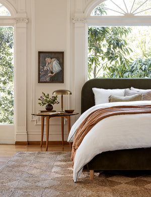 The Solene Loden Velvet platform bed sits atop a jute rug with a woven diamond pattern next to a portrait of a woman