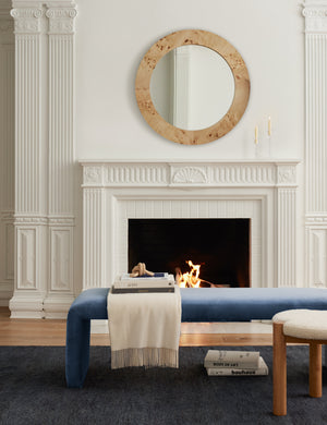 The Tate navy velvet upholstered bench sits in a living room with a burl wood mirror hanging over a fireplace and a cashmere throw blanket with books sitting on top of it.