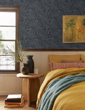 #color::pink #insert::down #insert::polyester | The Vadala long lumbar pink patterned pillow lays atop a bed with mustard-toned linens in a bedroom with blue patterned walls and a sculptural wooden nightstand