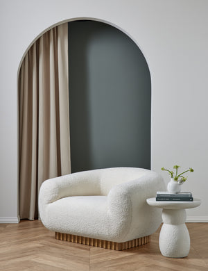 The Billow ivory boucle lounge chair sits in a studio room atop chevron wooden floors and next to a rounded white side table