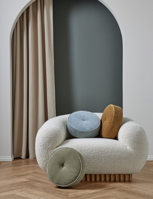 The Velvet Disc Pillow by Sarah Sherman Samuel in canyon blue, amber, and juniper green sit atop a boucle accent chair