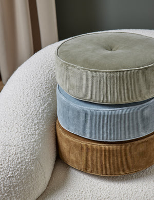 The Velvet Disc Pillow by Sarah Sherman Samuel in amber, canyon blue, and juniper green are stacked atop each other sitting on a white boucle accent chair