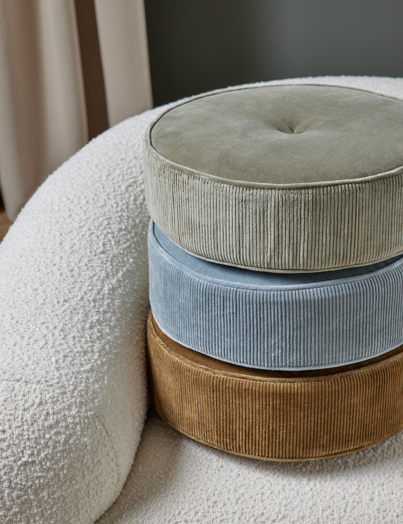 #color::canyon-blue | The Velvet Disc Pillow by Sarah Sherman Samuel in amber, canyon blue, and juniper green are stacked atop each other sitting on a white boucle accent chair