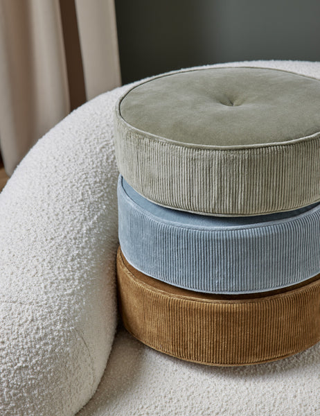 #color::canyon-blue | The Velvet Disc Pillow by Sarah Sherman Samuel in amber, canyon blue, and juniper green are stacked atop each other sitting on a white boucle accent chair