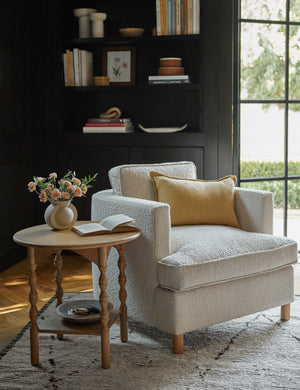 The arlo marigold lumbar pillow sits on an accent chair next to a sculptural wooden side table