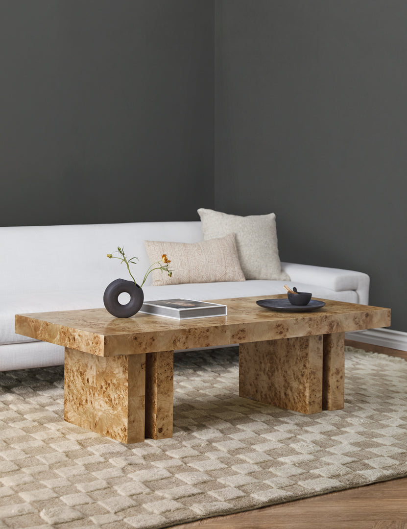 | The Brisa rectangular burl wood coffee table with four legs sits atop a light tan and ivory checkerboard area rug and in front of a white modern sofa.