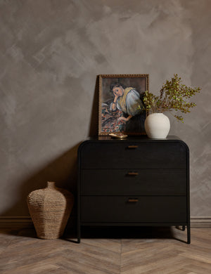 The Brooke 3-drawer black oak dresser sits against a gray wall next to a jute basket with a portrait and a white vase