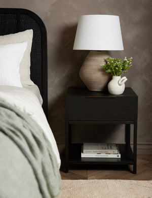 Dana Black Wood Nightstand sits underneath a ribbed table lamp and to the right of a bed with a black woven frame