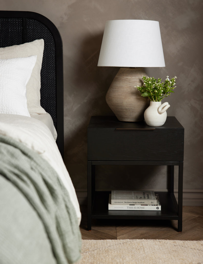 #color::black | Dana Black Wood Nightstand sits underneath a ribbed table lamp and to the right of a bed with a black woven frame 