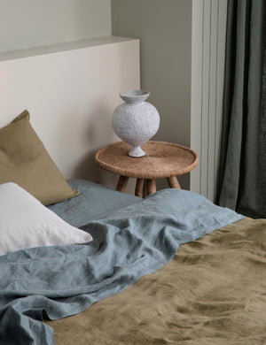 The European Flax Linen bluestone Sheet Set by Cultiver lays on a bed in a bedroom with olive green linens and a natural wooden nightstand
