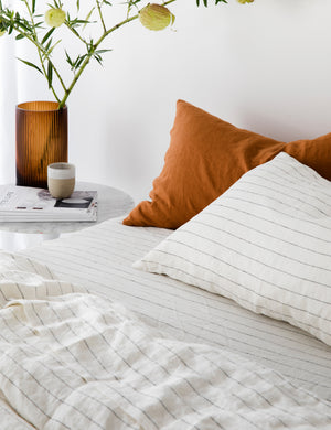 The European Flax Linen pencil stripe Sheet Set by Cultiver lays on a bed in a bedroom with cedar orange linens and a marble nightstand with an amber vase sitting atop it