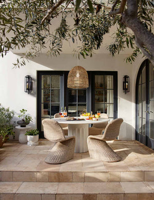 The Doric white round dining table sits on a patio underneath a woven chandelier surrounded by wicker dining chairs