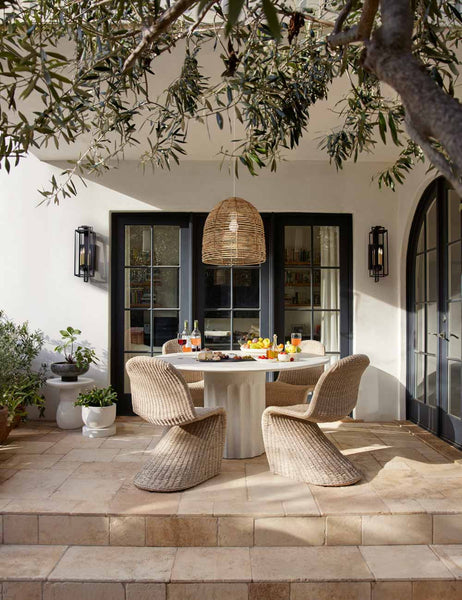 | The Doric white round dining table sits on a patio underneath a woven chandelier surrounded by wicker dining chairs