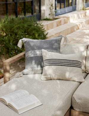 Marchesa agate gray indoor and outdoor square pillow with tasseled corners sits on a natural linen sofa in an outdoor space with a natural and black patterned throw pillow