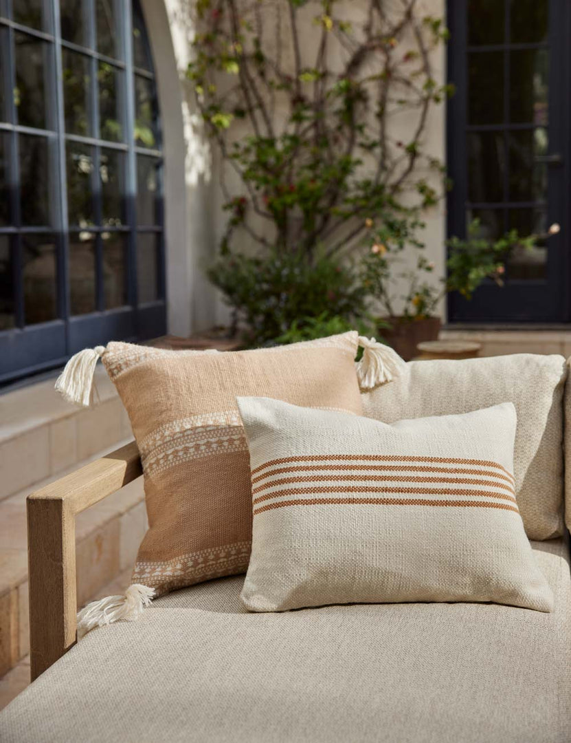 #color::sandstone #style::square | Marchesa sandstone indoor and outdoor square pillow with tasseled corners sits on a natural linen sofa with an orange striped throw pillow