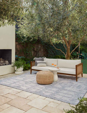 The Yamina blue indoor and outdoor rug lays in an outdoor space under a woven coffee table and a wood-framed sofa