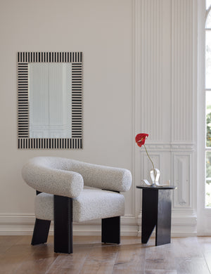 The Celeste black wood accent chair with wishbone frame sits in a bright room with a black wood side table, a black and white stripe framed mirror, and a sculptural candle holder.
