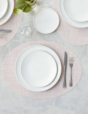 The Set of four blush pink Mini Basketweave Oval Placemat by Chilewich sits under a white dinnerware set