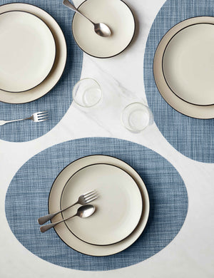 The Set of four chambray blue Mini Basketweave Oval Placemat by Chilewich sits under a cream dinnerware set