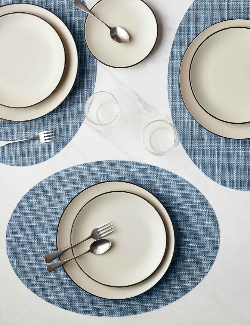 #color::chambray | The Set of four chambray blue Mini Basketweave Oval Placemat by Chilewich sits under a cream dinnerware set