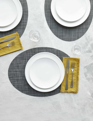 The Set of four cool gray Mini Basketweave Oval Placemat by Chilewich sits under a white dinnerware set
