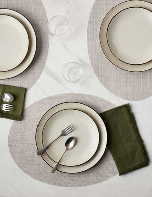 The Set of four parchment-toned Mini Basketweave Oval Placemat by Chilewich sits under a cream dinnerware set