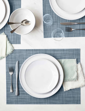 The Set of four chambray blue Mini Basketweave Rectangle Placemat by Chilewich sits under a white dinnerware set