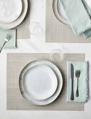 The Set of four parchment-toned Mini Basketweave Rectangle Placemat by Chilewich sits under a white dinnerware set