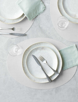 The Set of four sandstone gray Mini Basketweave Oval Placemat by Chilewich sits under a white speckled dinnerware set