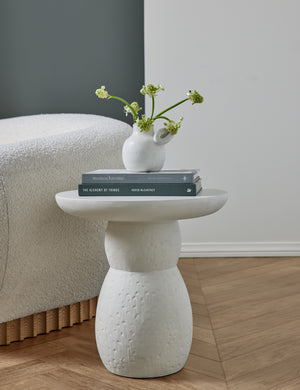 The Clouded White Round Side Table sits in a studio with a white vase and stack of books next to a white boucle accent chair