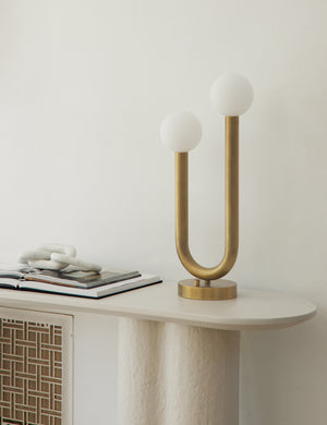 The Happy gold, natural brass table lamp by Regina Andrew with a dual-metal tube silhouette with contrasting matte white bumbs sits on a white sideboard next to marble decor and a stack of open books