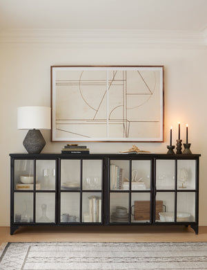 The Marjorie black iron sideboard with glass paneled doors is full of books, glasses and other decor items, and has three lit tapered candles and a dark gray lamp on it while an abstract neutral painting hangs above it.