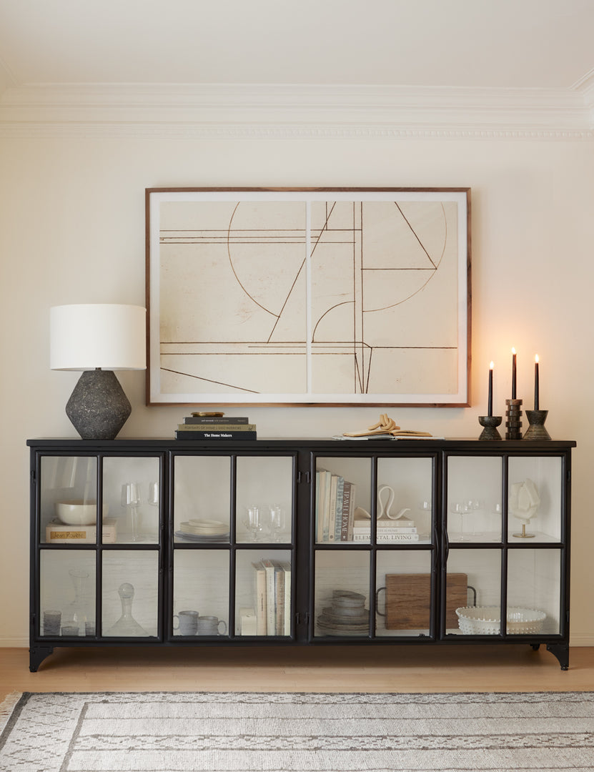 | The Marjorie black iron sideboard with glass paneled doors is full of books, glasses and other decor items, and has three lit tapered candles and a dark gray lamp on it while an abstract neutral painting hangs above it.