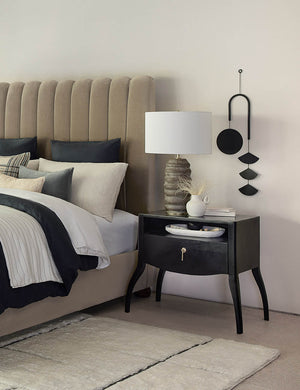 The Ola earth-toned ridged Ceramic Table Lamp by Regina Andrew sits atop a black wooden nightstand in a bedroom with a velvet framed bed