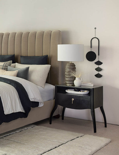 | The Ola earth-toned ridged Ceramic Table Lamp by Regina Andrew sits atop a black wooden nightstand in a bedroom with a velvet framed bed