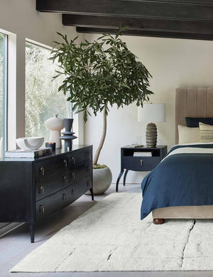 The Anabella black wood dresser with silver drawer pulls sits in a bedroom underneath two sculptural vases with a gray velvet bed and anabella nightstand to the right of it.