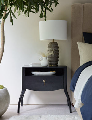 The Anabella black wood nightstand has a lamp with a ribbed base, a stack of books, and a white vase sitting atop it