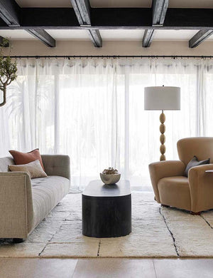 The costa rug lays in a living room with a large window and sheer curtains under a beige velvet accent chair and linen sofa