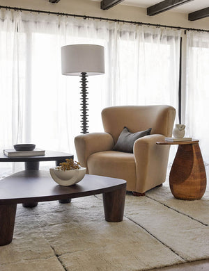 The Fishbone black ebony Floor Lamp by Regina Andrew with a gradated wooden base and linen drum shade sits in a living room with nested coffee tables, a beige velvet accent chair, and a wooden textured side table